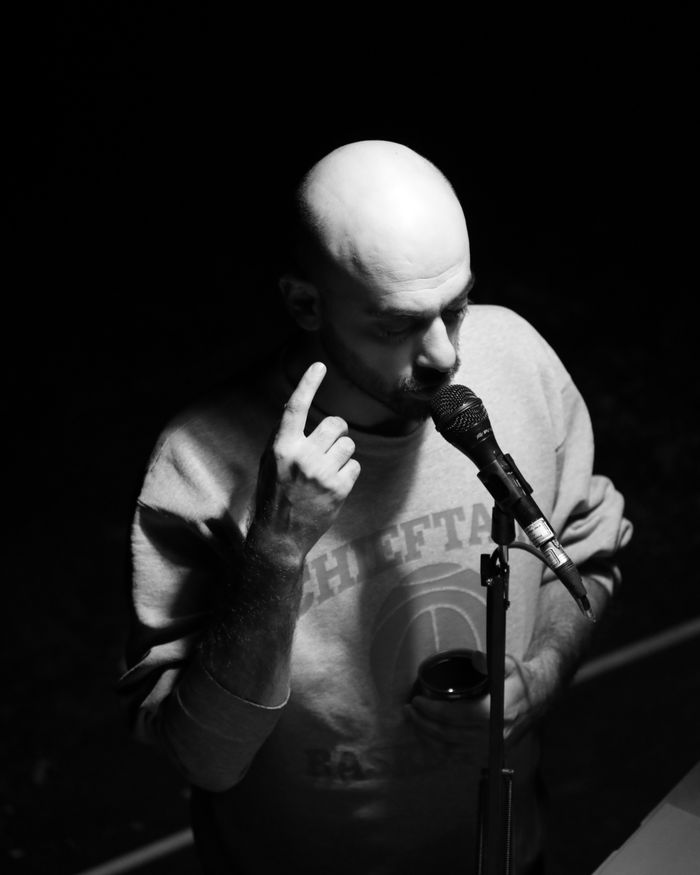 Bald adult man performing with a microphone in an artistic black and white setting.